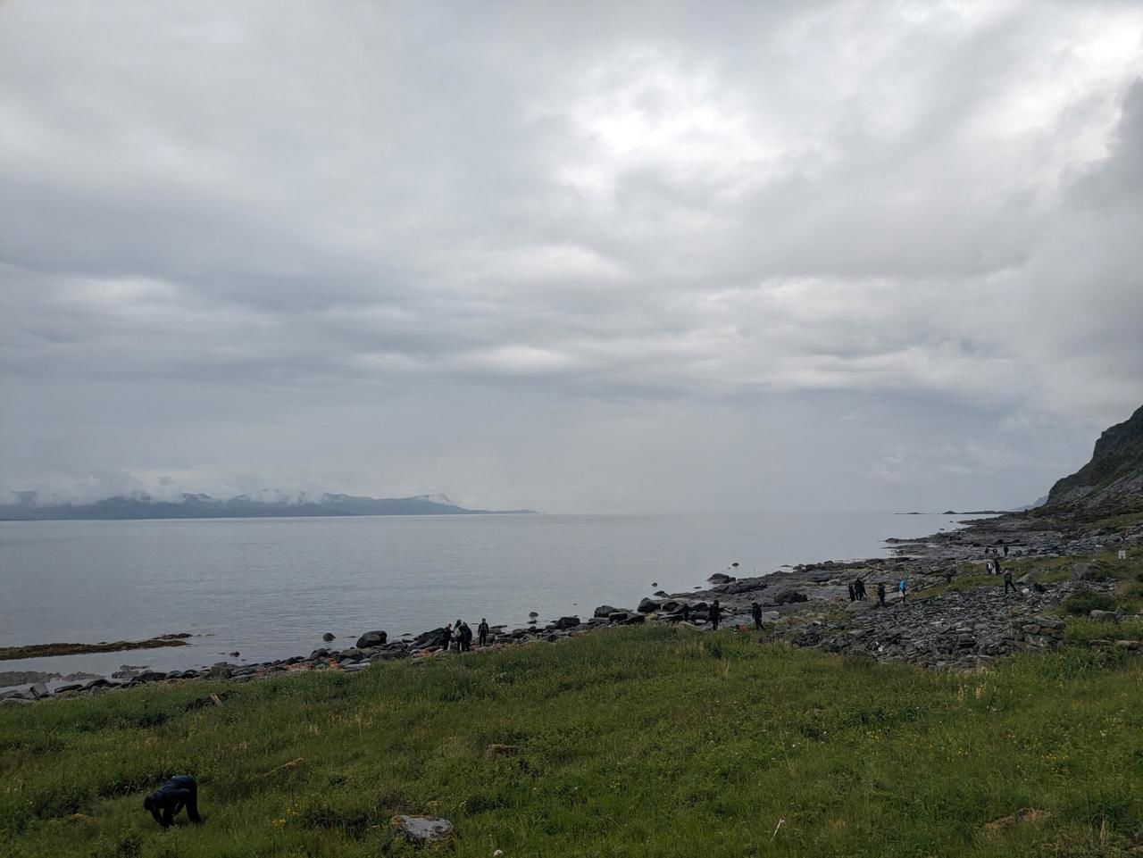 Landskapsbilete med elevar i bakgrunnen. Dei er langt vekke. Grønt gras i framgrunnen og hav i bakgrunnen.
