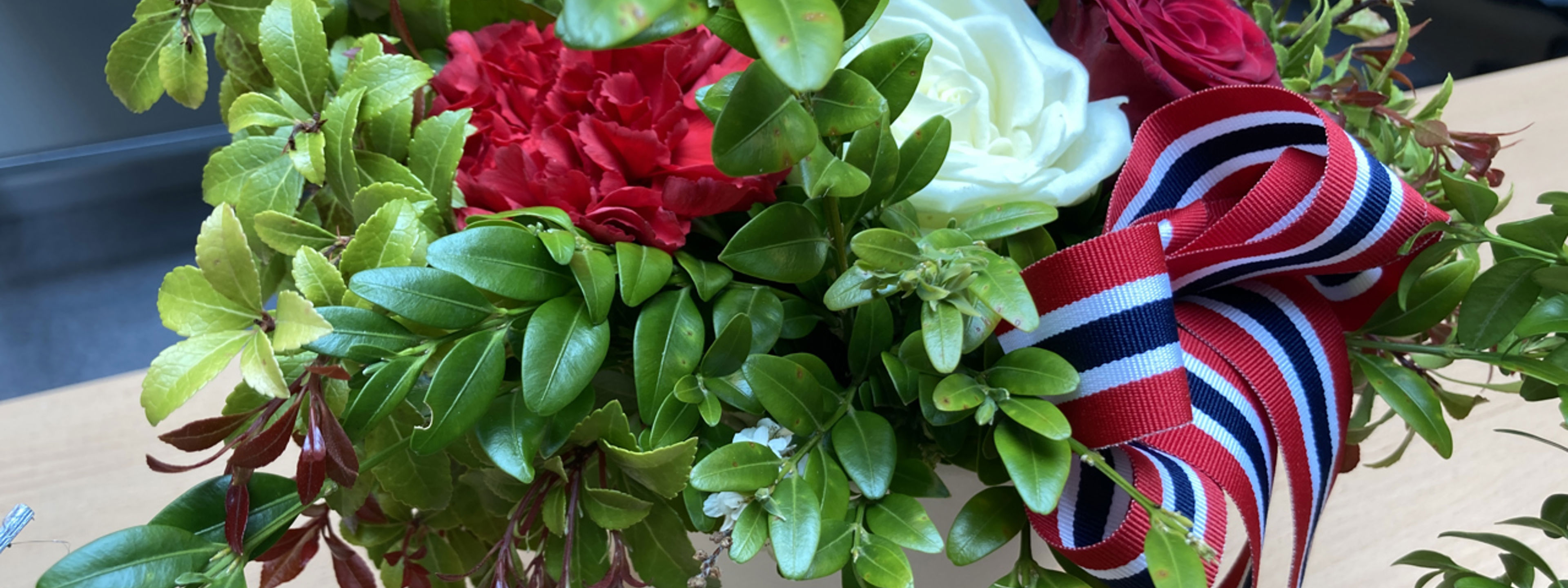Bomsterdekorasjon med kvit rose, raud rose, band i dei norske fargane og ein del grønt.