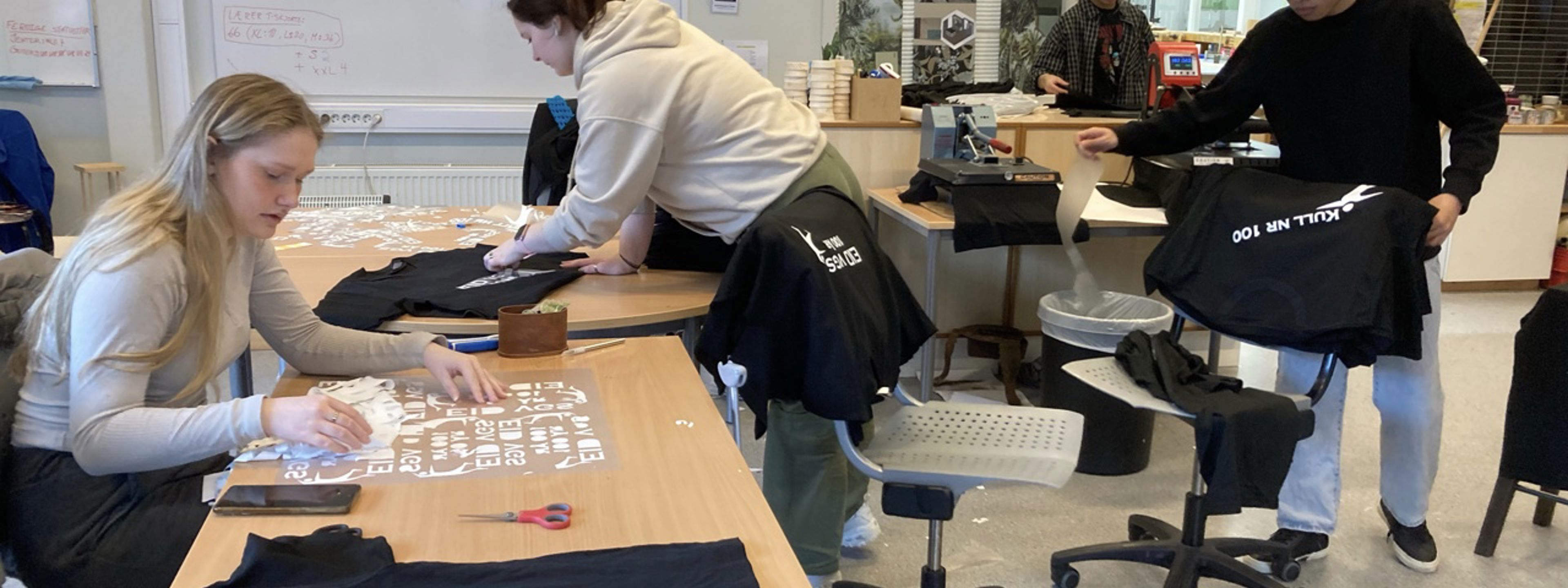 Fire elevar på eit klasserom som produsera t-skjorter.  Dei står og sit ved eit bord. Nokon strippa og nokon pressar.